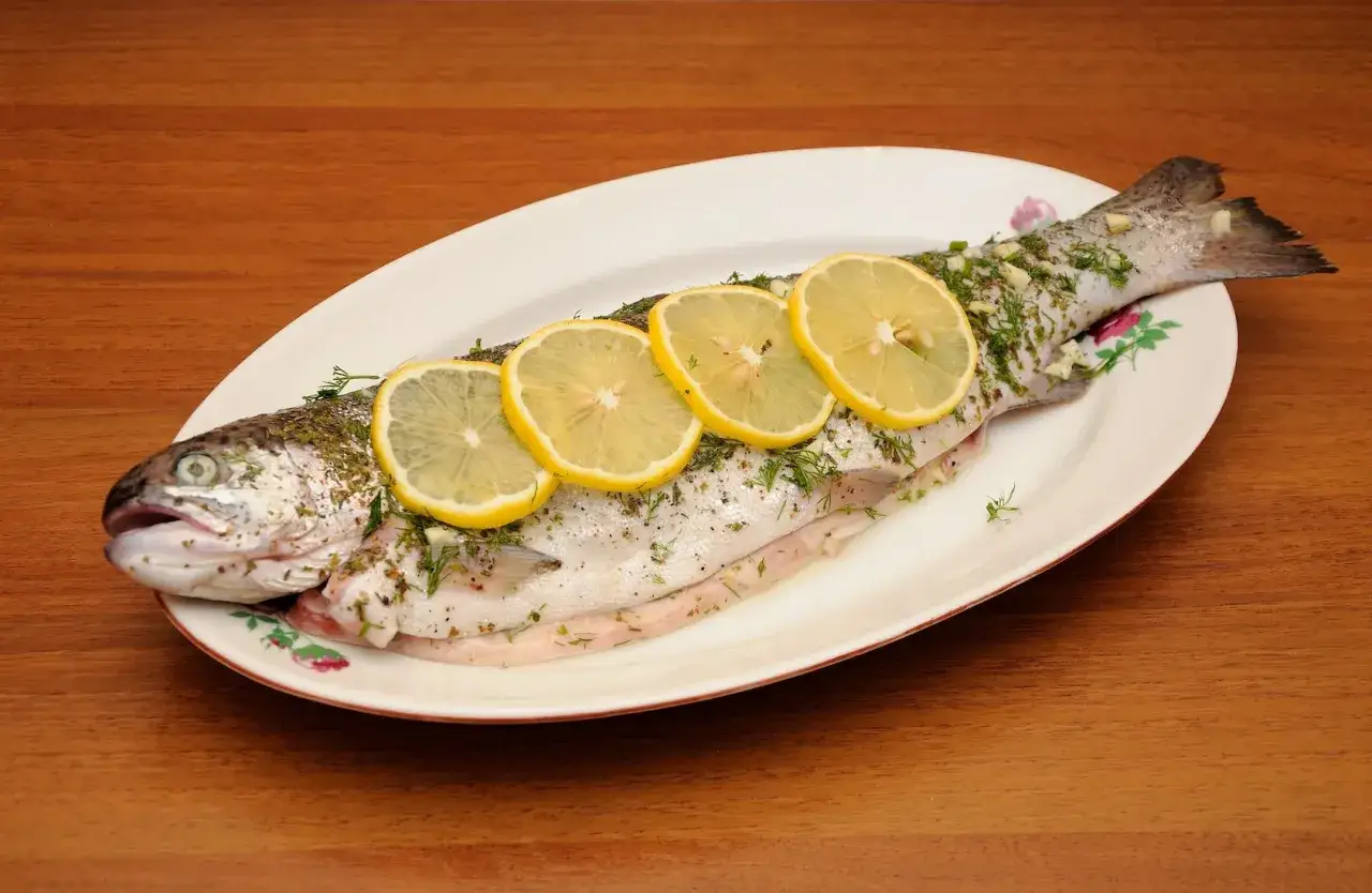 A whole trout, seasoned with herbs and garlic, is garnished with lemon slices on a white oval plate.