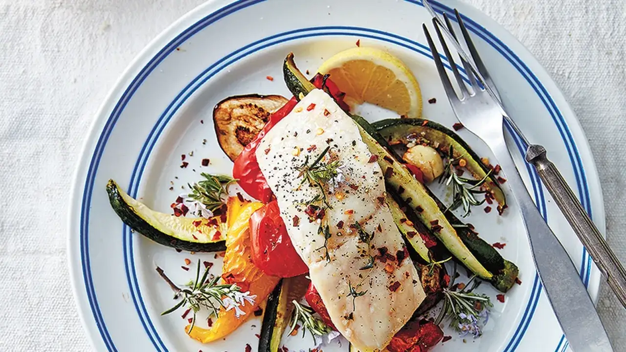 A delicious grouper recipe featuring a white fish fillet atop roasted zucchini, eggplant, bell peppers, and tomatoes, seasoned with herbs and chili flakes.