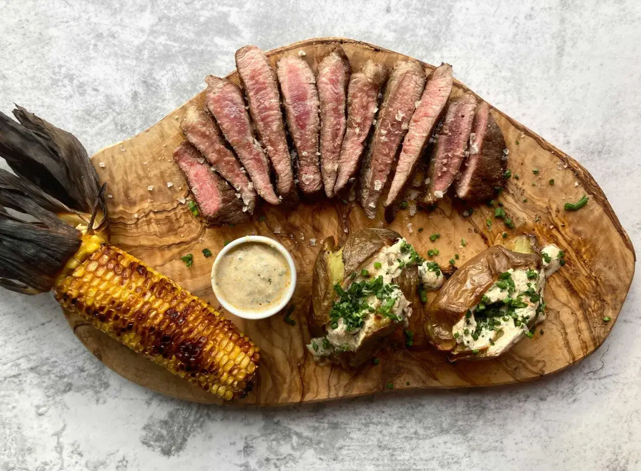 Juicy pork presa, grilled corn, and loaded baked potatoes served on a wooden board.