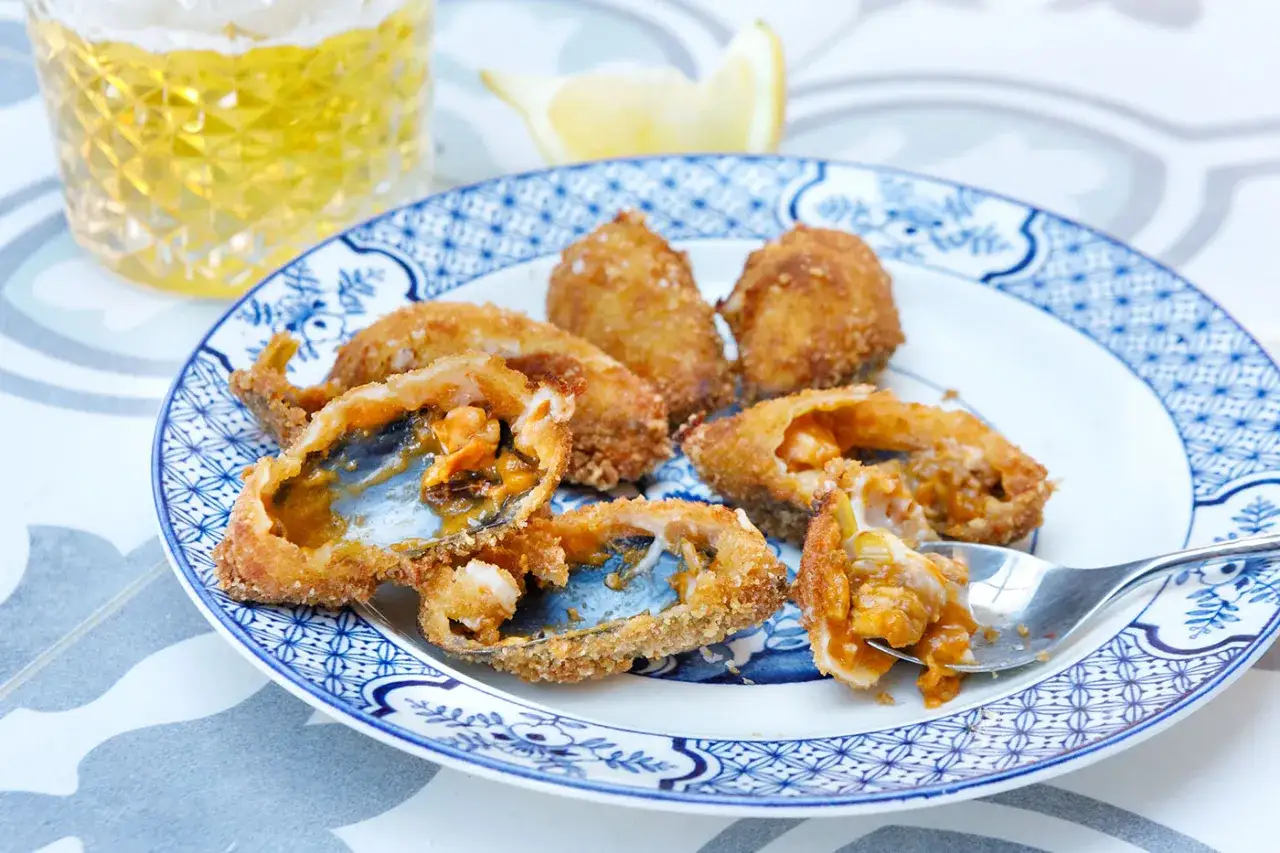 Crispy fried tiger mussels, some split open to reveal the succulent interior, sit on a blue and white patterned plate next to a glass of beer.