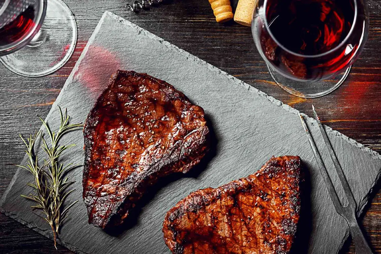 Two grilled steaks on a slate board, with glasses of red wine, a rosemary sprig, and wine accessories. A perfect pairing for merlot vs cabernet.