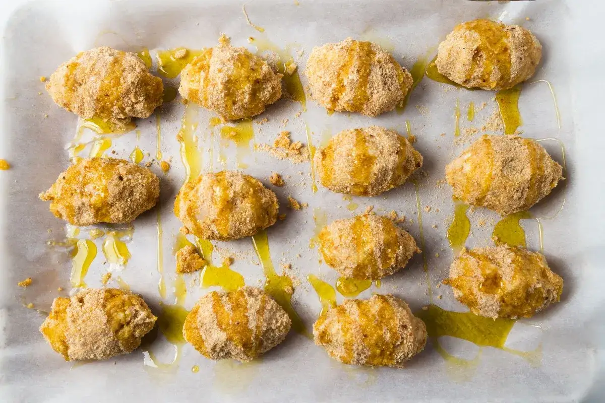 Twelve breaded croquetas de pollo drizzled with olive oil on parchment paper, ready for baking.
