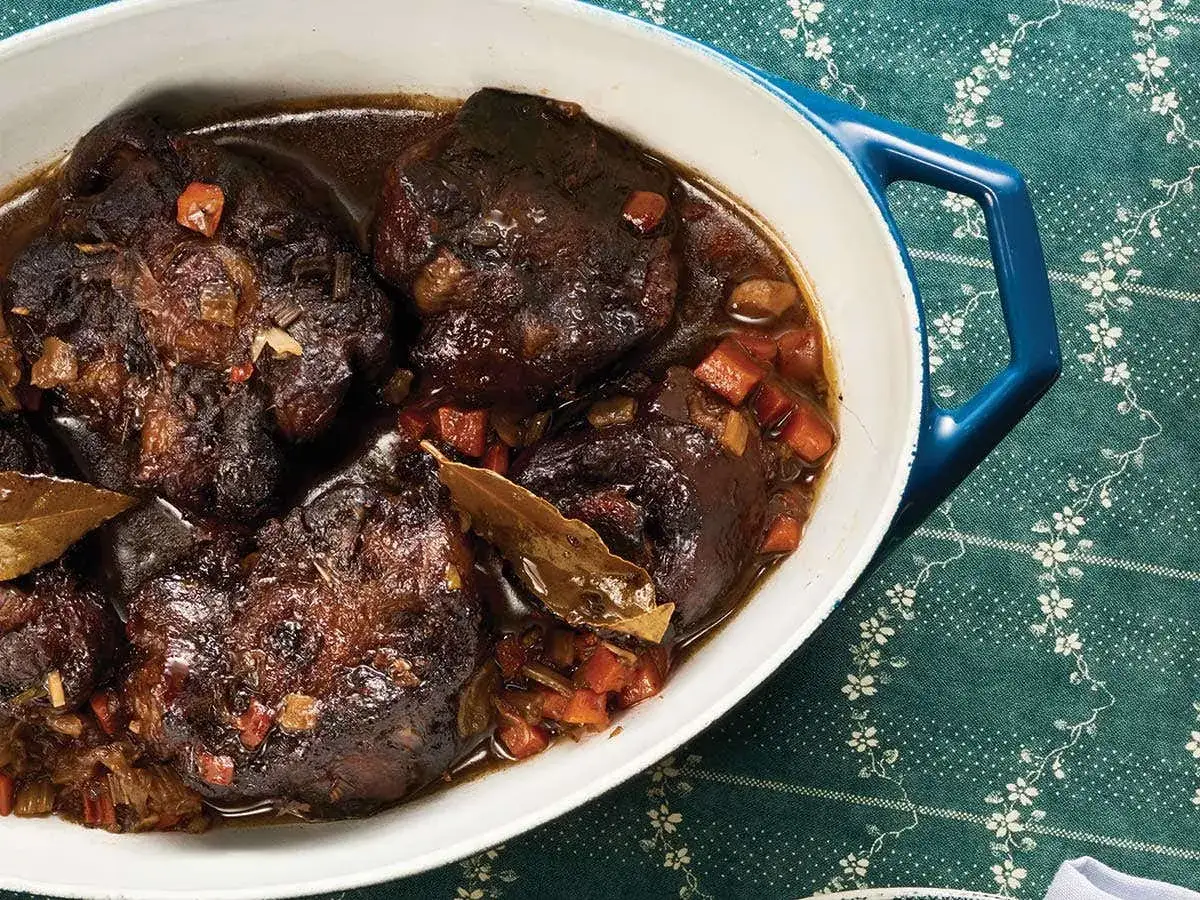 A rich oxtail recipe in a white oval dish, braised with carrots and bay leaves on a floral green tablecloth.