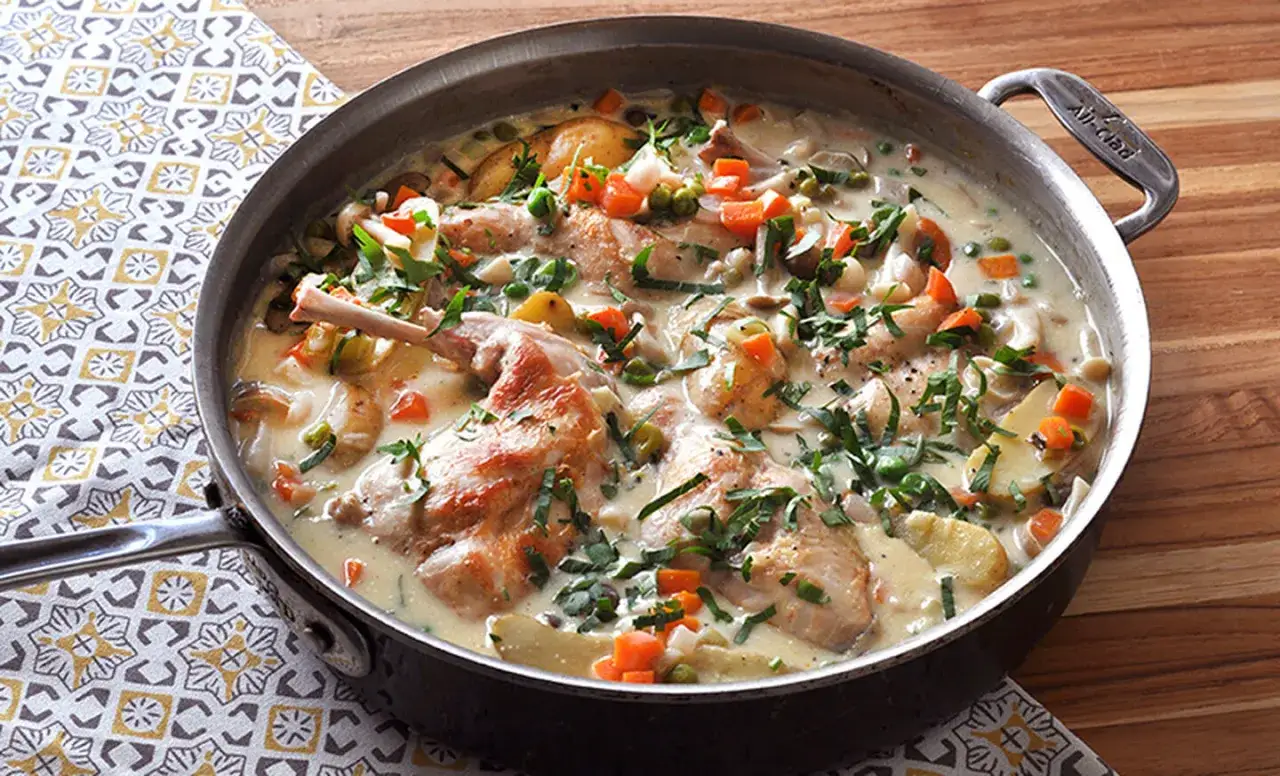 A hearty rabbit stew simmers in a large pan, filled with tender rabbit pieces, potatoes, carrots, peas, and herbs in a creamy sauce.