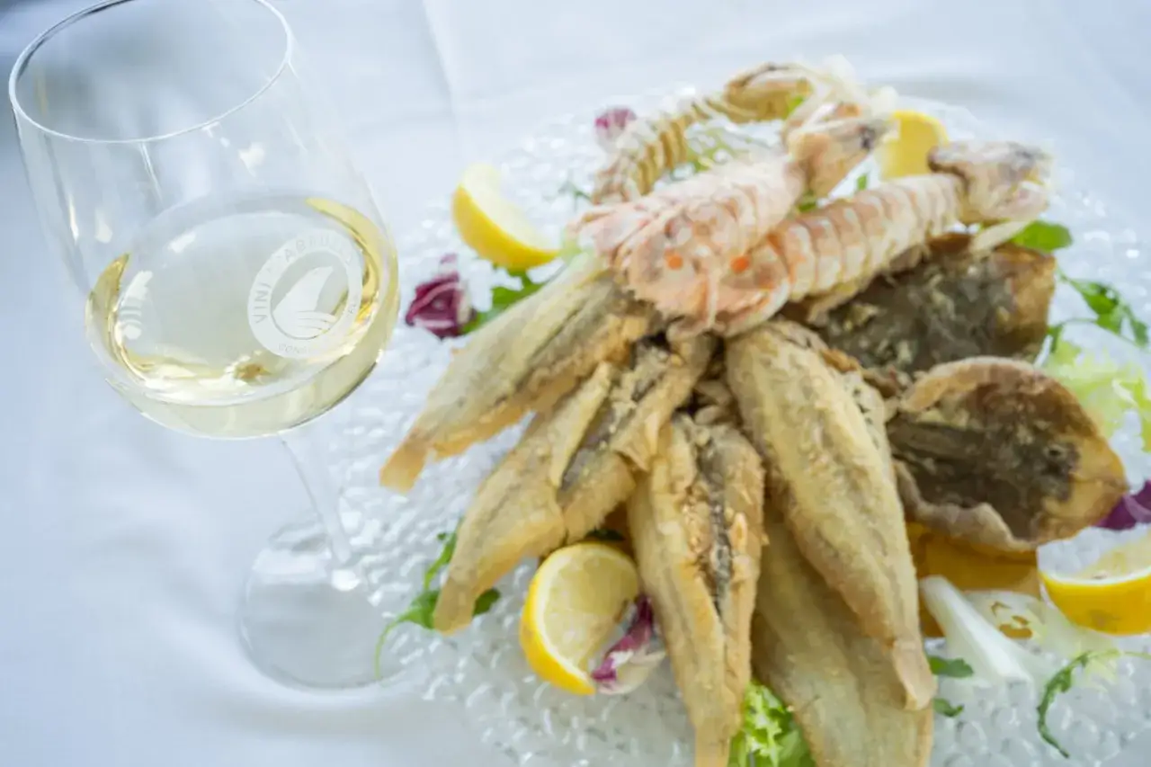 A glass of trebbiano wine sits beside a platter of fried seafood, including small fish and mantis shrimp, garnished with lemon and greens.
