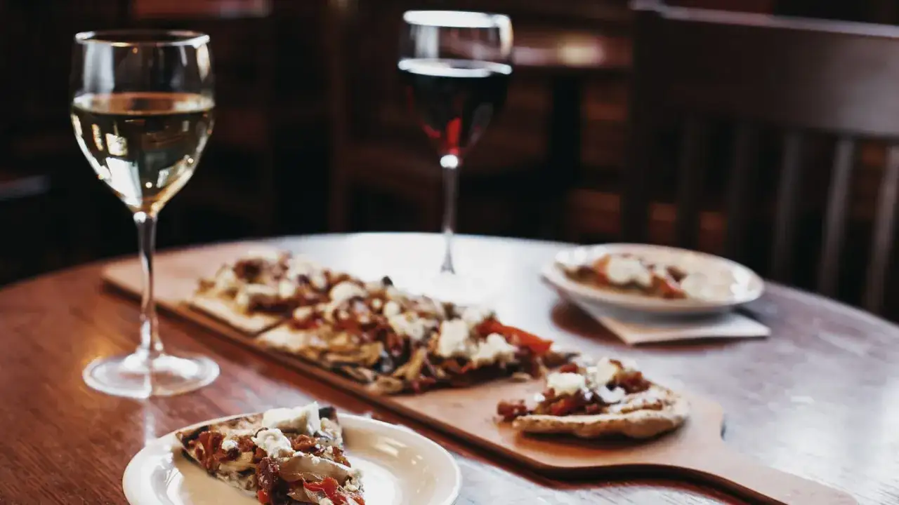 A glass of white gamay wine sits beside a wooden board with flatbread pizzas and a slice on a plate.