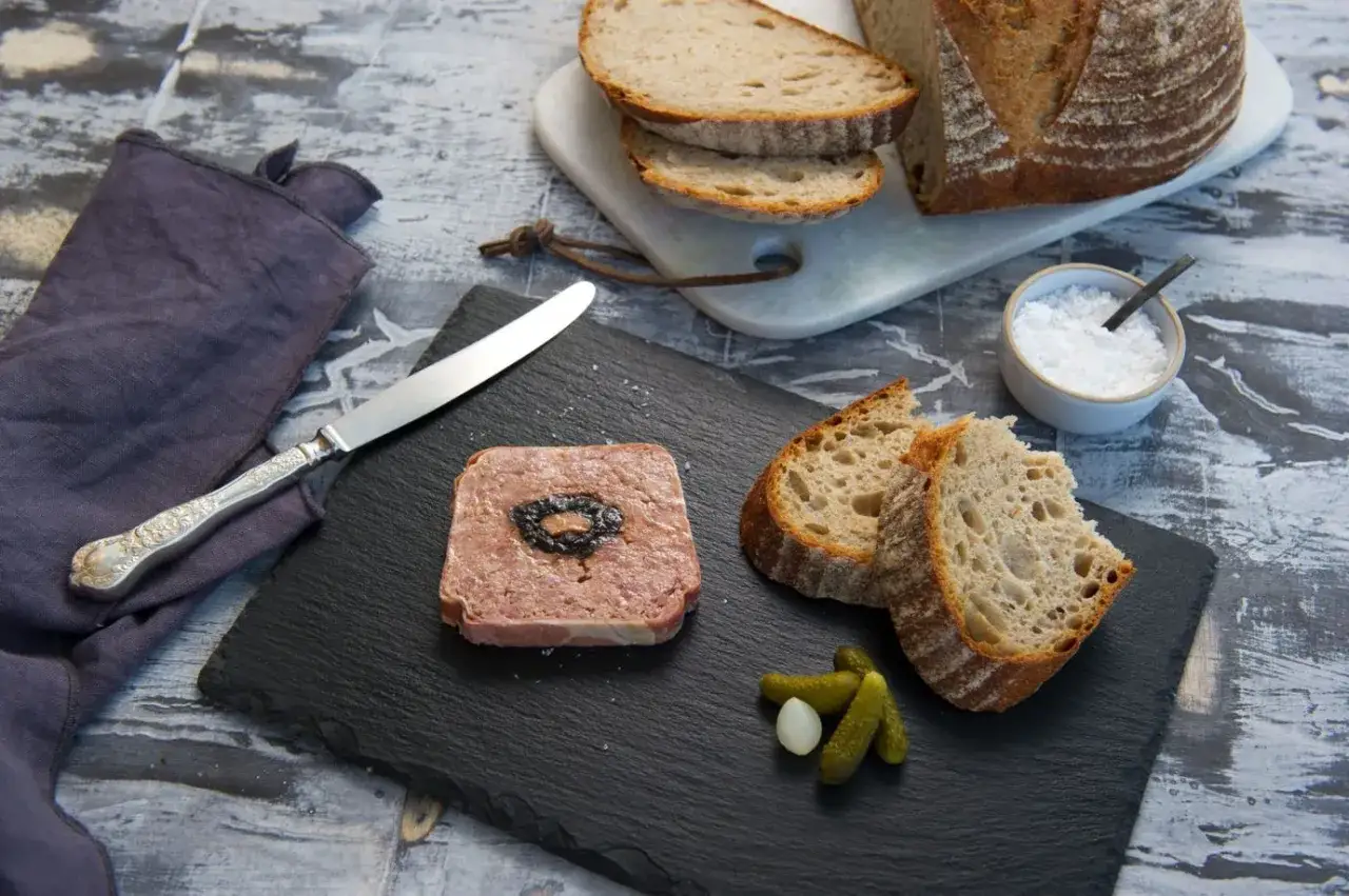A square of duck terrine with a dark center sits on a slate board next to crusty bread slices, cornichons, and a small dish of salt.