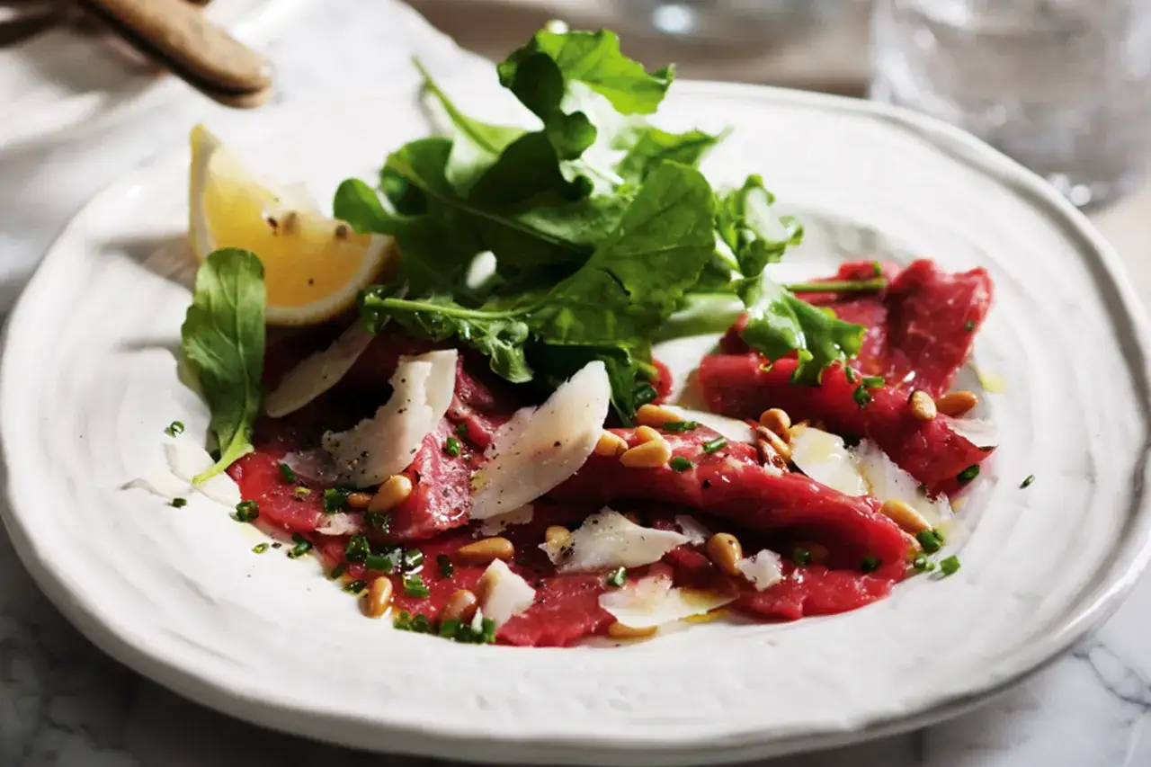 Thinly sliced beef carpaccio with arugula, pine nuts, shaved Parmesan, and a lemon wedge.