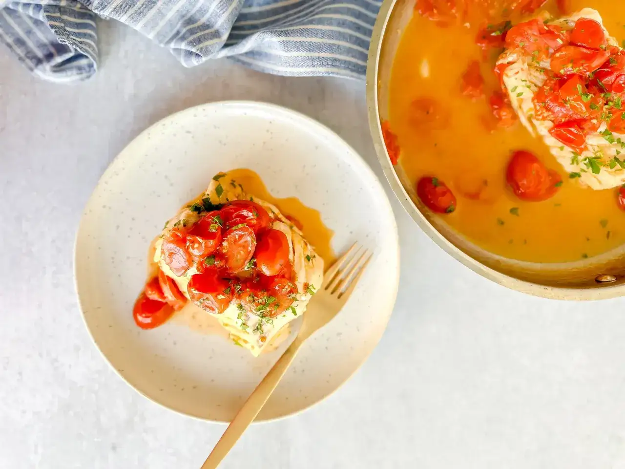 A plate with a piece of poached fish topped with cherry tomatoes and parsley, next to a pan with more fish and sauce. Delicious poached fish recipes!