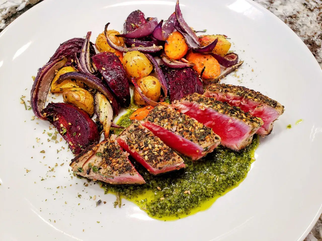 Seared tuna tataki, coated in sesame seeds and herbs, served with roasted root vegetables and a vibrant green sauce.