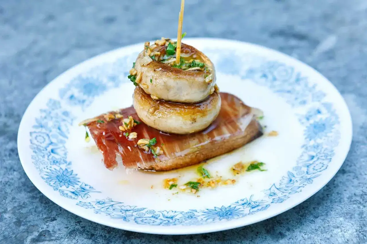 A delicious grilled mushroom recipe featuring two stacked mushrooms on a slice of prosciutto, drizzled with garlic and herbs.