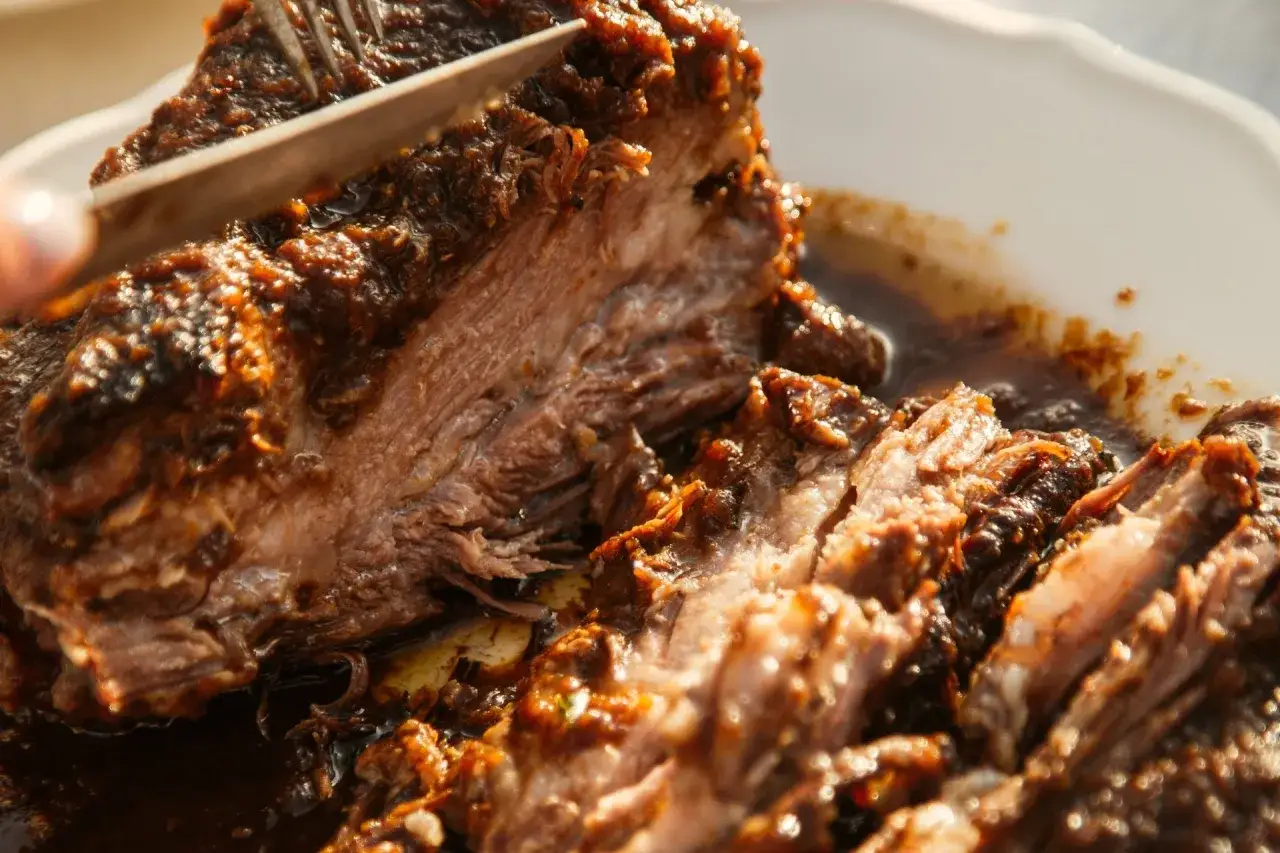 A fork and knife are slicing into a tender, juicy iberico pork shoulder recipe, glistening with a rich, dark sauce.