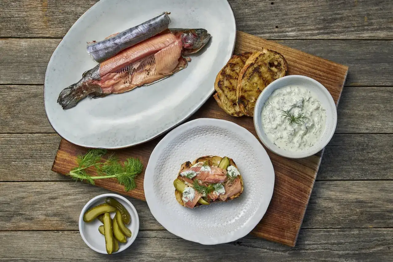 A whole smoked trout is presented alongside grilled bread, dill sauce, pickles, and a slice of toast topped with smoked trout, pickles, and dill.