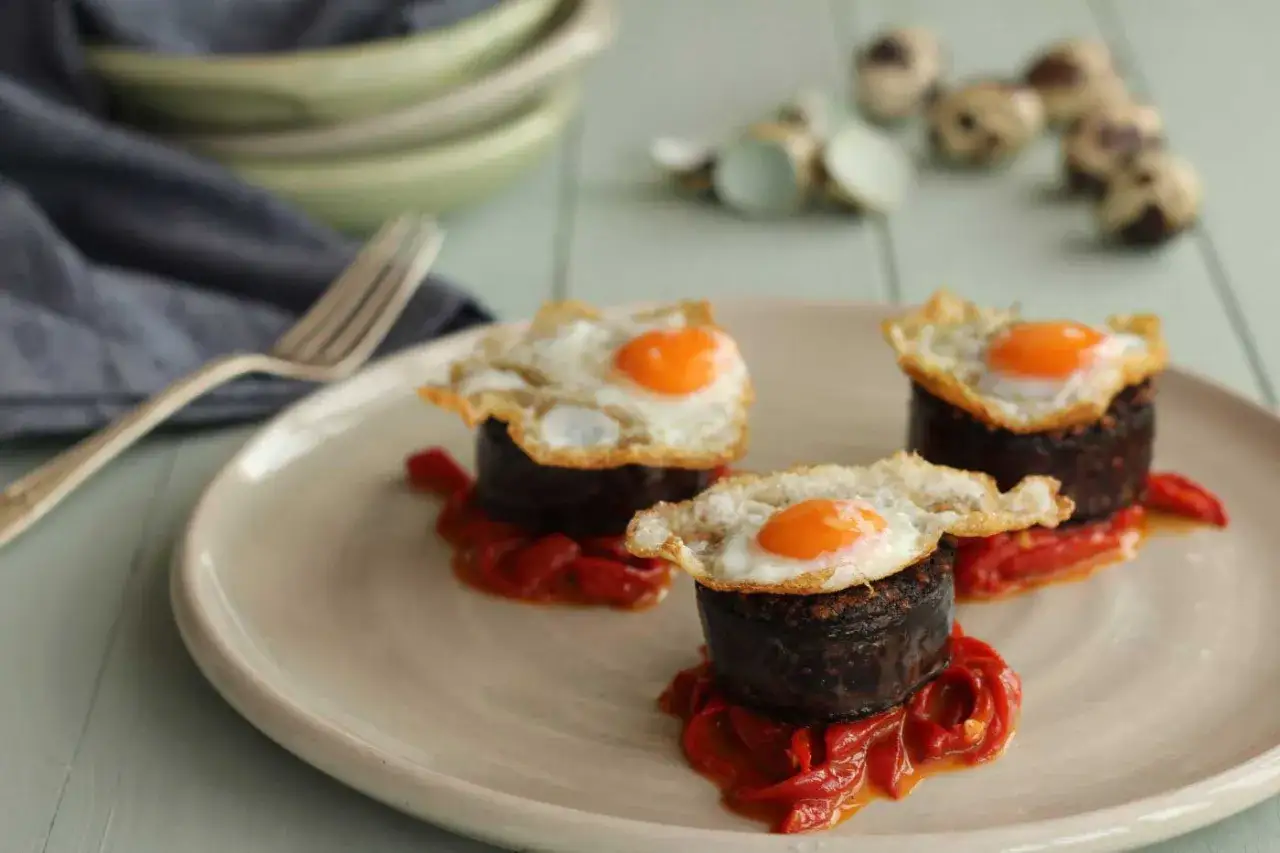 Three morcilla pintxos, each a dark cylinder topped with a fried quail egg, rest on a bed of red peppers on a light plate.
