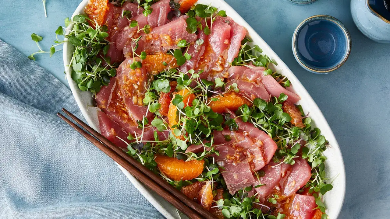 Tuna crudo, thinly sliced raw tuna, is artfully arranged with vibrant microgreens and citrus segments, drizzled with a light dressing.