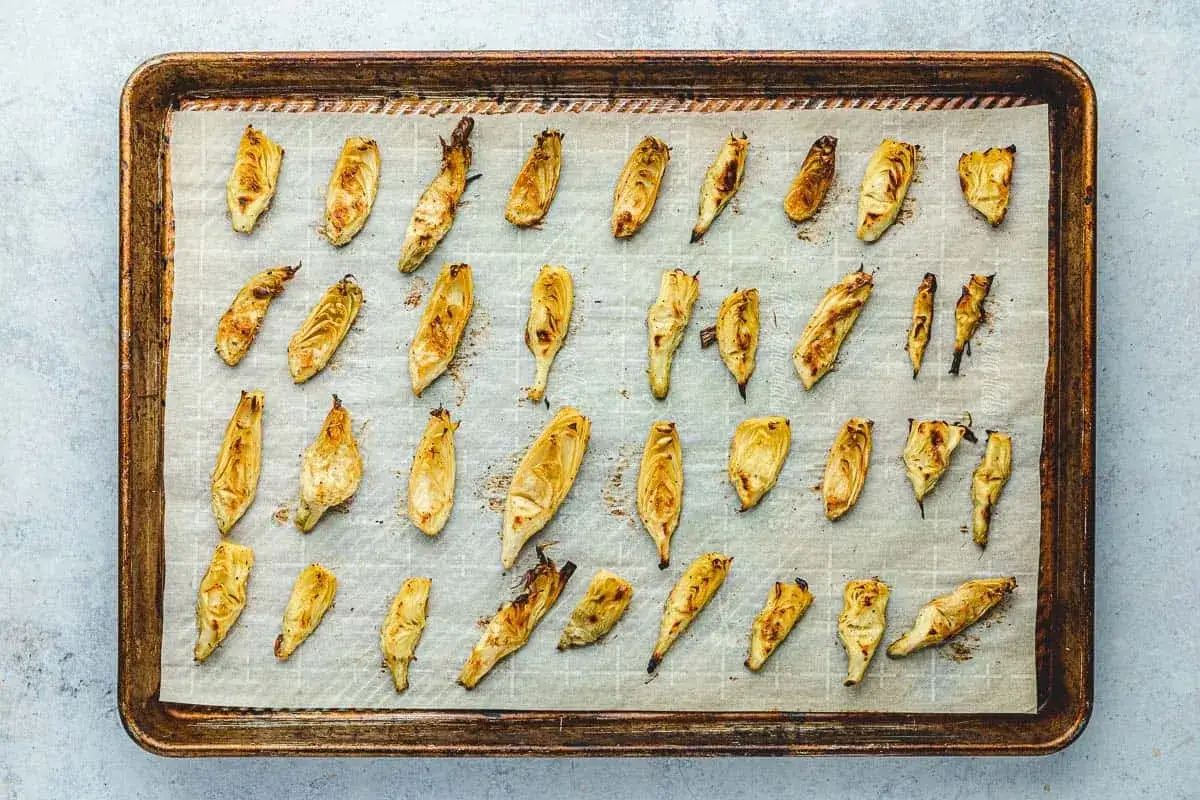 Roasted artichoke hearts, golden brown and slightly charred, are arranged on a baking sheet lined with parchment paper, ready to be pickled.