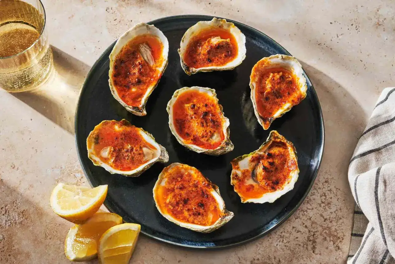 Seven baked oysters topped with a spicy red sauce, ready to be enjoyed as part of delicious smoked oyster recipes. Served with lemon wedges and a glass of white wine.