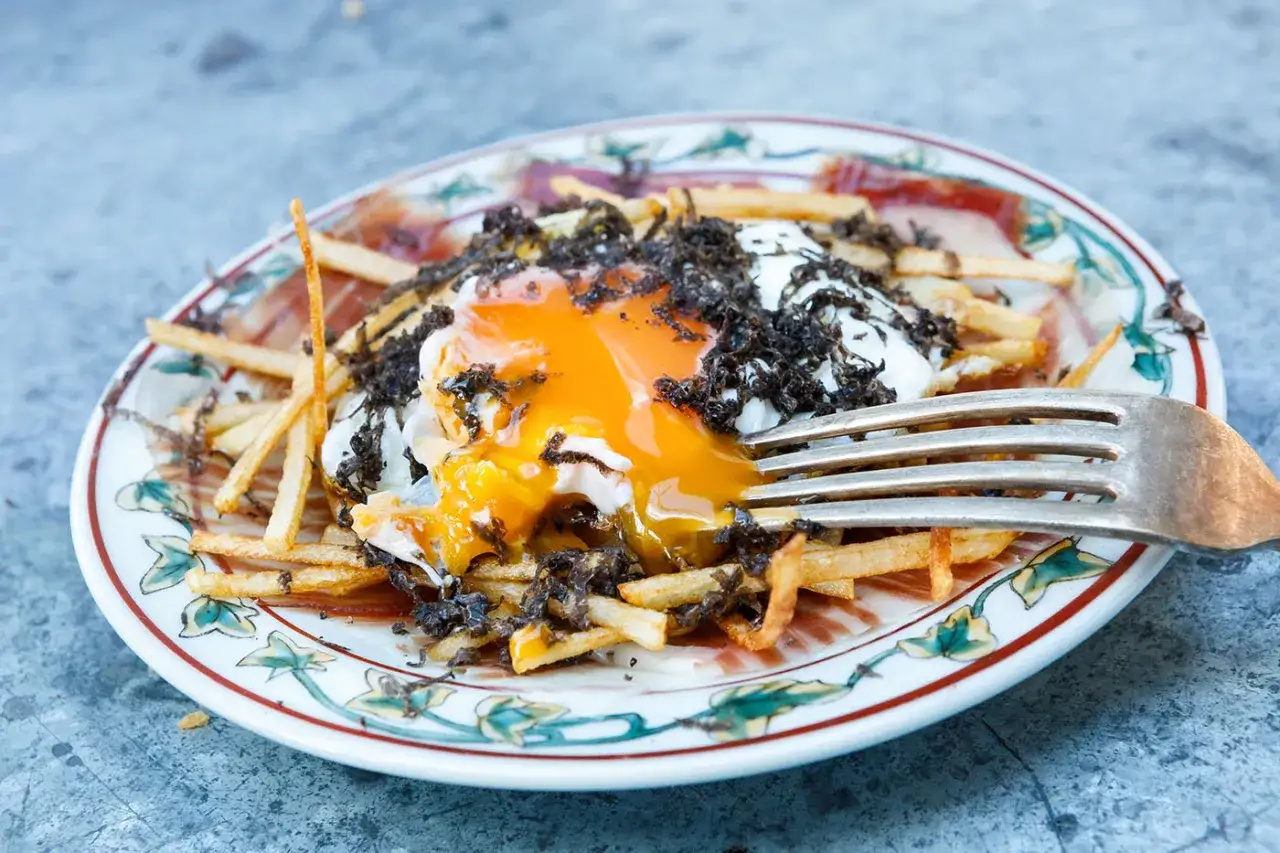 A fork pierces a runny egg yolk atop crispy fries and black truffle shavings, a delicious plate of huevos rotos.