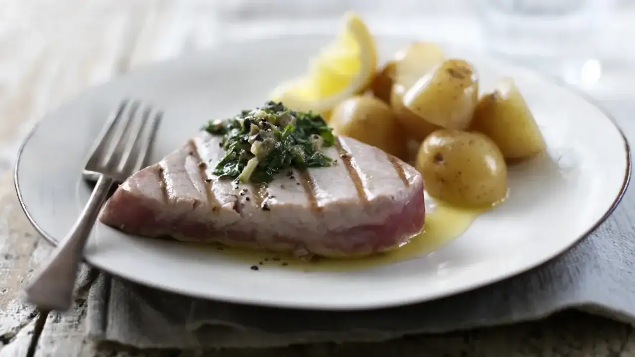 A grilled tuna steak, topped with a fresh herb sauce, sits beside a pile of baby potatoes and a lemon wedge. This atun food is a delicious and healthy meal.