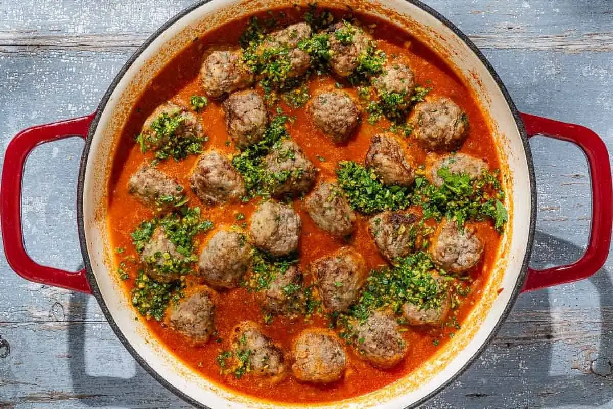 A close-up of a red-handled pan filled with delicious albondigas españolas, simmered in a rich tomato sauce and sprinkled with fresh parsley.