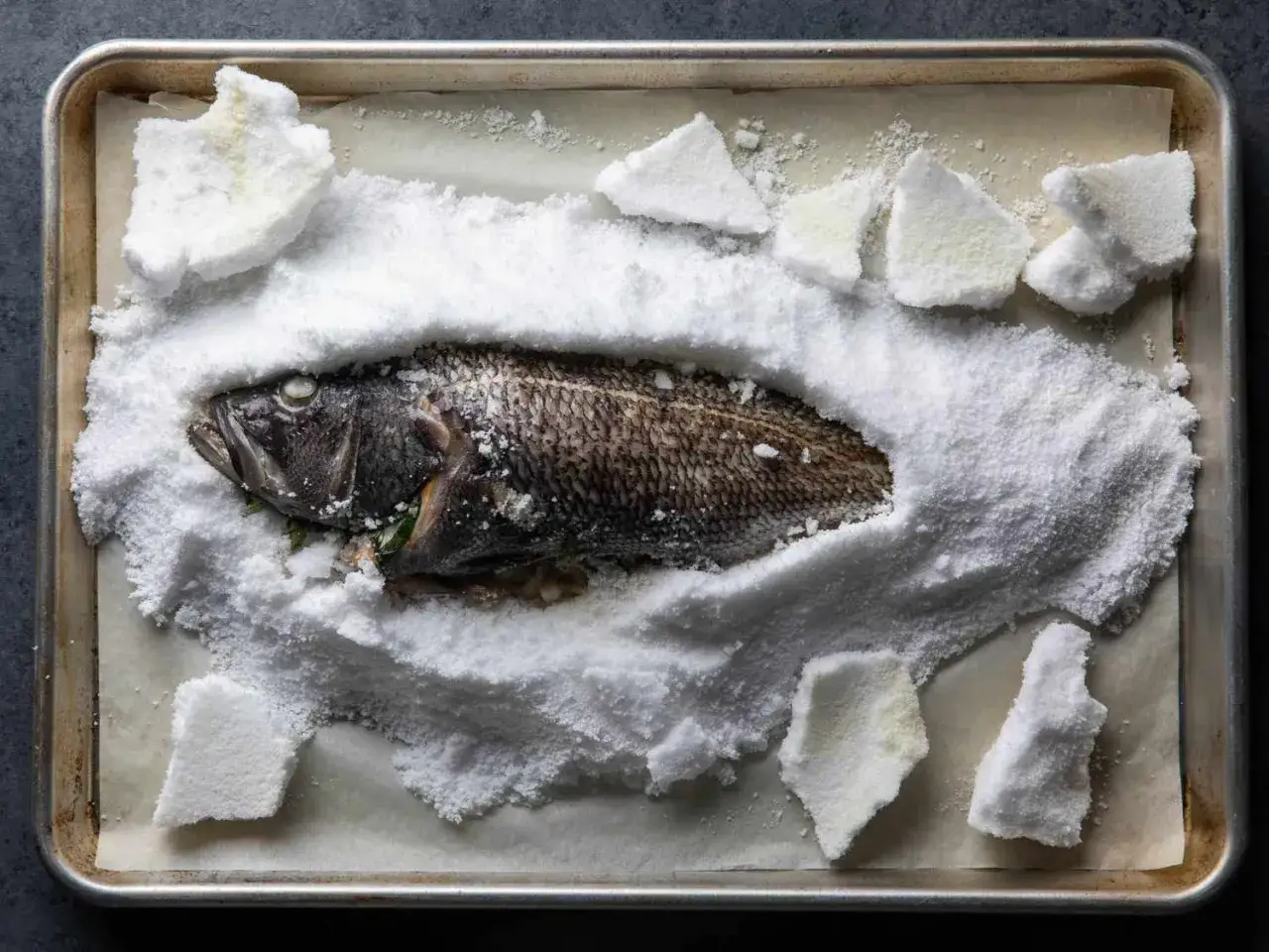 A whole salt crusted sea bass rests on a baking sheet, its scales visible beneath a thick layer of salt.