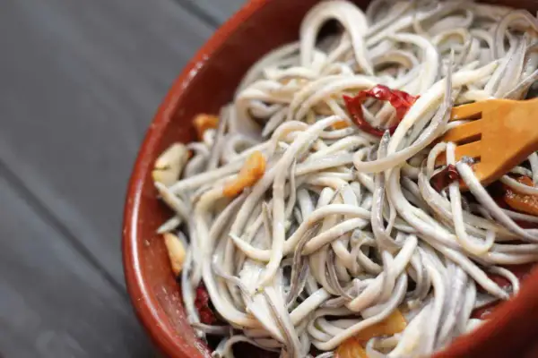 A close-up of a rustic bowl filled with a tangle of little eel, glistening with garlic and chili flakes. A wooden fork is nestled within the dish.