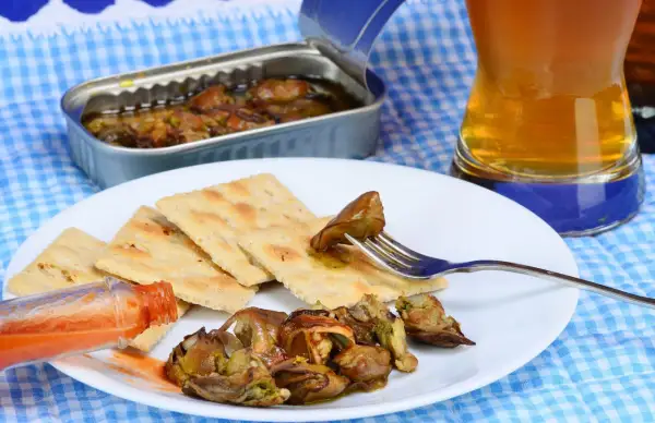 A fork lifts a smoked oyster from a plate of crackers, a bottle of hot sauce and a beer nearby. Delicious smoked oyster recipes are easy to make with these ingredients.