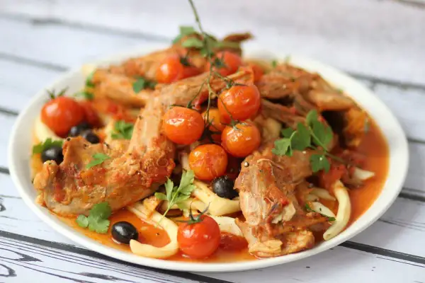 A hearty rabbit stew with tender meat, cherry tomatoes, olives, and pasta, garnished with fresh parsley.