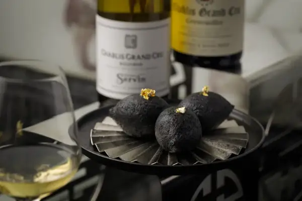 A glass of chablis white wine sits beside a plate of three dark, spherical treats topped with gold leaf, with bottles of chablis grand cru in the background.