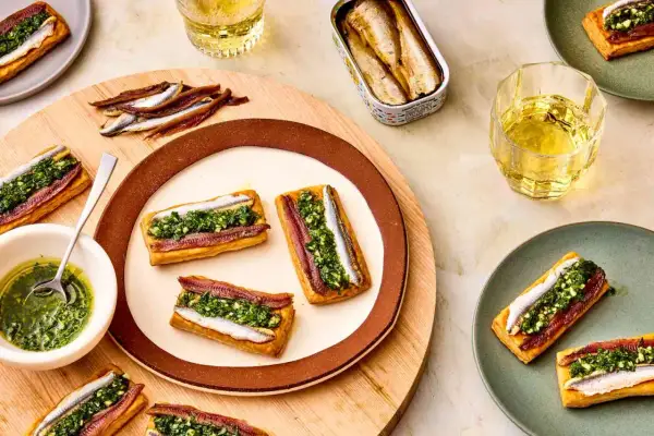 Rectangular pastries topped with red and white anchovies and green sauce. A tin of white anchovies sits nearby.