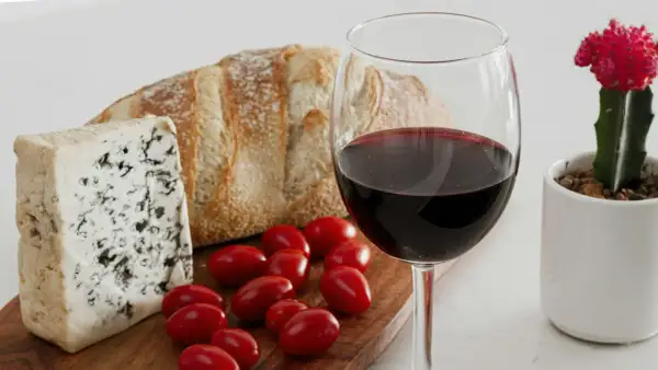 A glass of red wine, perhaps a bold trebbiano wine, sits beside a rustic loaf of bread, blue cheese, and cherry tomatoes, with a small potted cactus nearby.