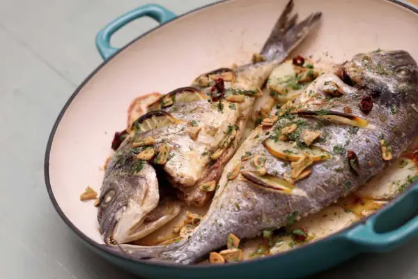 Two whole sea bream baked with lemon slices, garlic, and herbs in a teal pan, served over sliced potatoes.