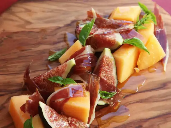 A refreshing melon con jamon appetizer with fresh figs and basil, drizzled with honey on a wooden board.