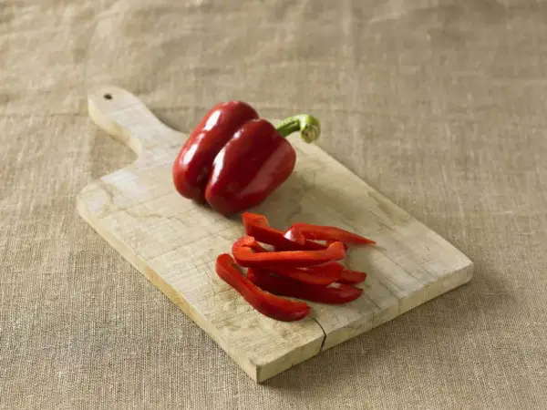 Red bell peppers, one whole and some sliced, on a wooden cutting board. Perfect for learning how to preserve peppers for later use.