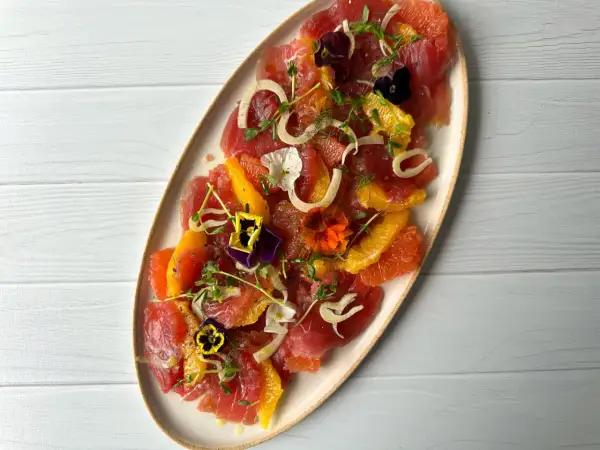 A vibrant tuna crudo dish featuring thinly sliced raw tuna, citrus segments, fennel, microgreens, and edible flowers.