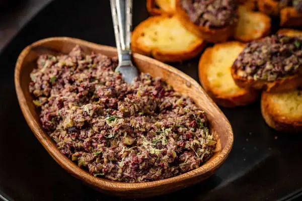 A rustic wooden bowl overflows with chunky tapenade, a savory spread of olives and herbs, ready to be enjoyed with toasted crostini.