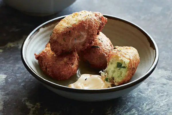 Golden-brown croquetas de bacalao, one broken open to reveal a fluffy interior, sit in a rustic bowl with a dollop of creamy sauce.