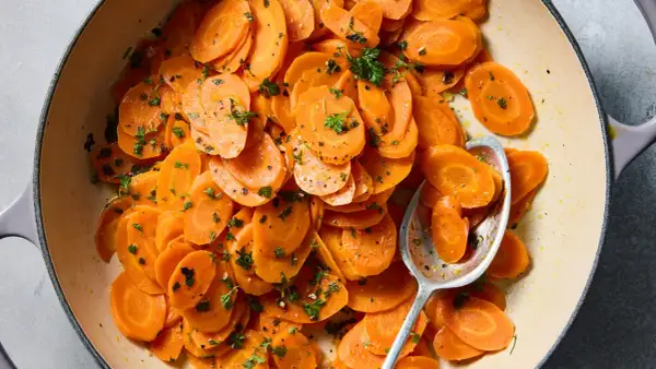 A close-up of a pot of glazed vichy carrots, glistening with butter and sprinkled with fresh parsley. A spoon rests in the pot, ready to serve.