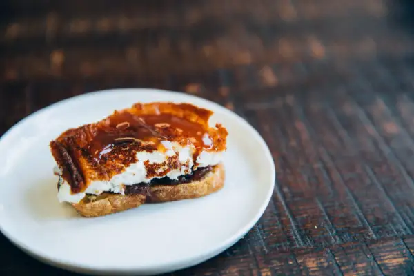 A decadent goat cheese toast, caramelized and drizzled with a rich, glossy sauce, sits on a white plate.