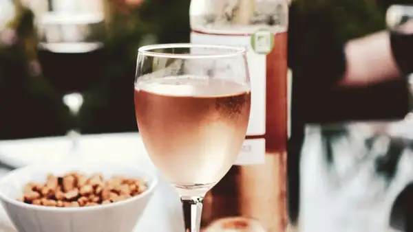 A glass of rosé wine, perhaps a Marsanne blend, sits on a table with a bowl of nuts and a bottle of wine.