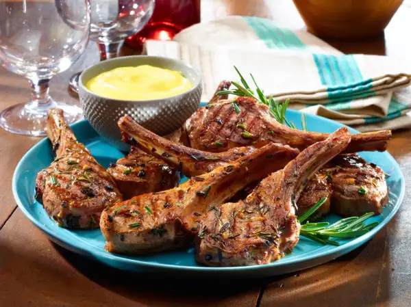 Grilled lamb chops, seasoned with rosemary, served on a blue plate with a side of yellow sauce. A taste of Spanish lamb meat.