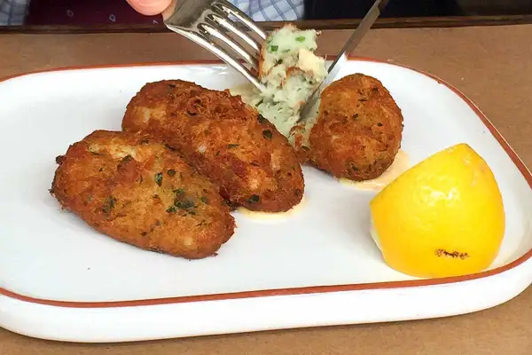 Golden brown cod fritters served with a dollop of mashed potatoes and a lemon wedge. A fork is lifting a bite of the fritter.