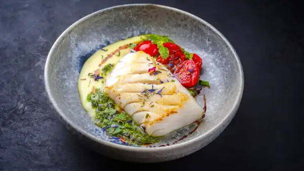 A beautifully plated poached fish recipe, featuring a white fish fillet with grilled marks, served with creamy sauce, pesto, and cherry tomatoes.