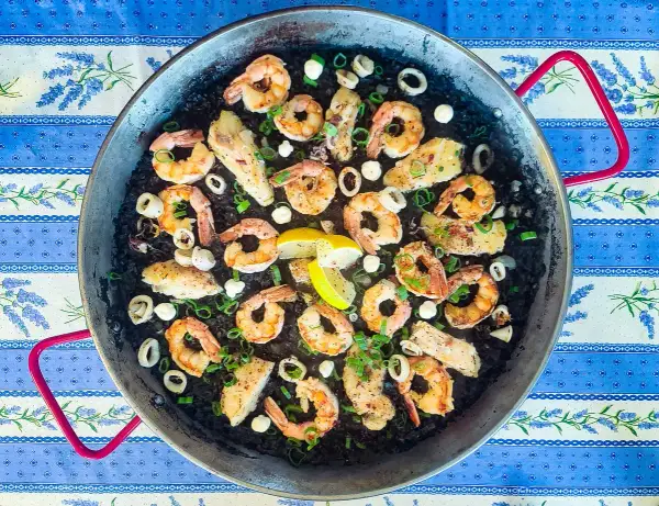 A delicious squid ink rice paella, brimming with plump shrimp, tender calamari rings, and flaky white fish, all garnished with fresh green onions and lemon wedges.