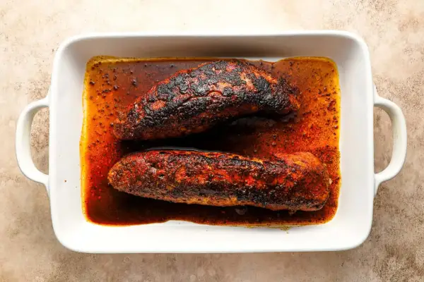 Two seasoned pork tenderloins, ready for a delicious solomillo recipe, rest in a white baking dish filled with rich, spiced juices.