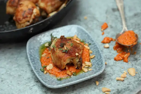 A succulent piece of chicken, part of a delicious chicken tapas dish, rests on a bed of vibrant orange sauce, garnished with herbs and chopped nuts.