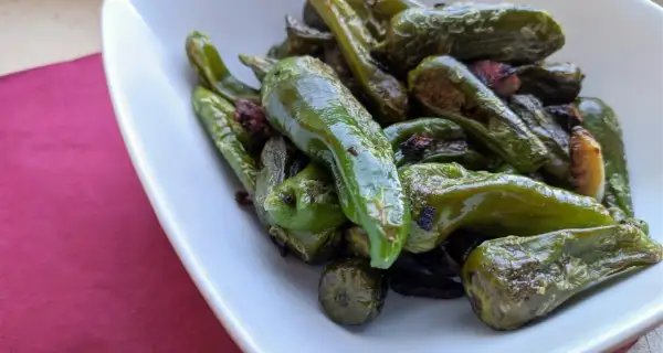 A pile of glistening, fried padron peppers in a white bowl, some with slightly charred edges, hinting at a delicious, savory snack.