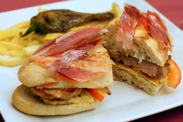 A delicious bocadillo with tender meat, cured ham, and fresh tomato, served with crispy fries and a roasted pepper.