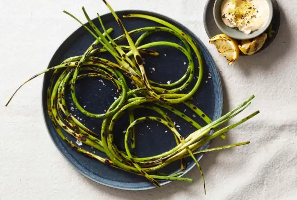 Grilled garlic scapes, seasoned with sea salt, are artfully arranged on a dark plate, with a small bowl of creamy dip and lemon wedges nearby.