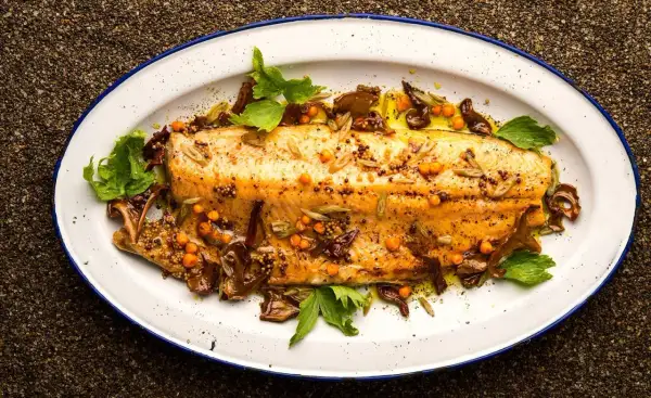 A beautifully broiled trout, glistening with olive oil and garnished with mustard seeds, sea buckthorn berries, and fresh herbs on a white enamel platter.
